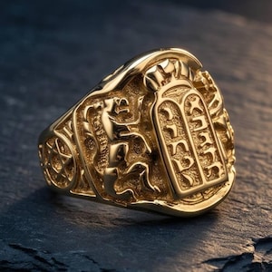 May include: A gold-toned ring featuring a raised relief design of the Ten Commandments tablets with Hebrew lettering, flanked by a lion figure on one side and a Star of David symbol on the other, all set within a textured, metallic band.