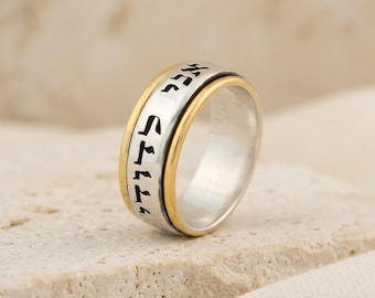 Könnte beinhalten: Ein zweifarbiger Spinner-Ring mit einem silbernen Mittelband, das schwarze hebräische Schriftzeichen trägt, und zwei goldfarbenen Außenrändern. Der Ring hat eine polierte metallische Oberfläche und ein breites, flaches Profil mit drehbarem Mittelteil.