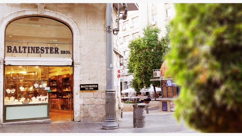 May include: A storefront with the sign "Balitnester Bros." above the entrance. The store appears to sell jewelry and other items. The storefront is located on a street with a lamppost and a tree in the background.