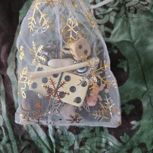 May include: A small, sheer white gift bag with gold snowflake accents, tied with a white ribbon. The bag contains various game pieces, including dice and small, light-colored game tokens. The background is a green and black patterned fabric.