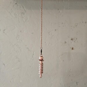 May include: A copper-colored pendulum with a spiral design hangs from a thin chain. The pendulum is suspended from a wooden surface.