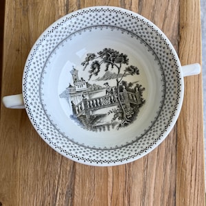 May include: White ceramic soup bowl with two handles, featuring a black and white landscape design inside. The rim has a decorative pattern. The bowl is set on a wooden surface.