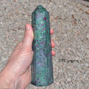 May include: A green and purple ruby zoisite crystal tower. The polished gemstone is a rectangular prism with a pointed top. The text on the image reads "597 grams".