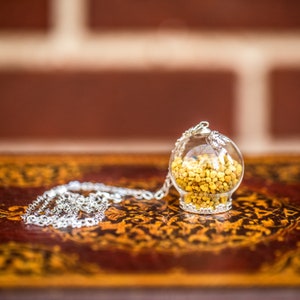 Puede incluir: Un collar de cadena de plata con un colgante de esfera de vidrio lleno de polen de abeja amarillo. El collar está sobre una superficie de madera con tallas intrincadas.