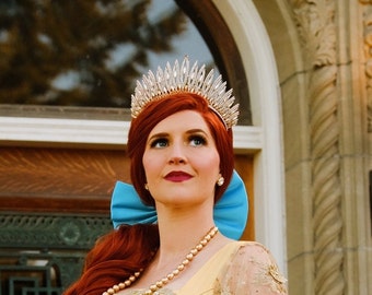 May include: A woman with long red hair wearing a gold crown, a blue bow, and a yellow dress with a gold necklace. She is standing in front of a building with a large window.