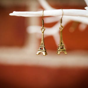 May include: A pair of gold-tone earrings featuring miniature Eiffel Tower charms. The earrings are hanging from a white branch.