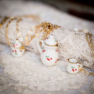 May include: A set of three miniature porcelain teapots with a cherry design. The teapots are on a white lace tablecloth and are connected by gold chains.