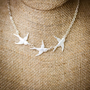 May include: Silver necklace featuring three silver bird charms. The necklace is displayed on a brown fabric mannequin.