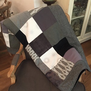 May include: A patchwork quilt with a gray, black, and white color scheme. The quilt is made of different fabrics and patterns, including polka dots, stars, and geometric shapes. The quilt is draped over a gray armchair with wooden legs.