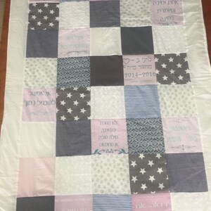 Puede incluir: Una colcha de retazos con una variedad de telas en tonos de rosa, gris y blanco. La colcha presenta una variedad de patrones, incluyendo estrellas, rayas y diseños florales. La colcha está personalizada con texto en hebreo.