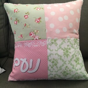 Peut inclure: Un coussin patchwork rose et vert avec des pois blancs et des motifs floraux. Le coussin porte le mot "עמי" écrit en blanc sur le devant.