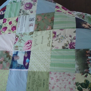 Op de afbeelding: Een patchworkdeken met verschillende bloemenpatronen en foto's. De deken is gemaakt van verschillende stoffen en kleuren, waaronder groen, blauw, roze en wit. De deken heeft een gepersonaliseerd bericht met de tekst "Shai Tocker" en "9/16/10".
