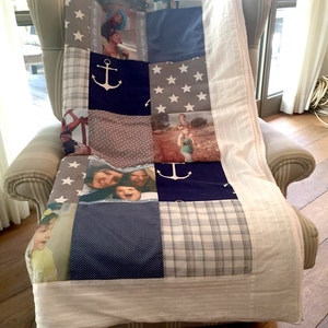 Op de afbeelding: Een wit en blauw patchwork-deken met een nautisch thema. Het deken heeft ankers, sterren en ruitjespatronen. Er zijn ook verschillende foto's van mensen op het deken.
