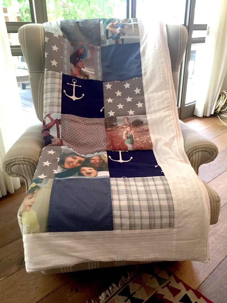 Op de afbeelding: Een wit en blauw patchwork-deken met een nautisch thema. Het deken heeft ankers, sterren en ruitjespatronen. Er zijn ook verschillende foto's van mensen op het deken.