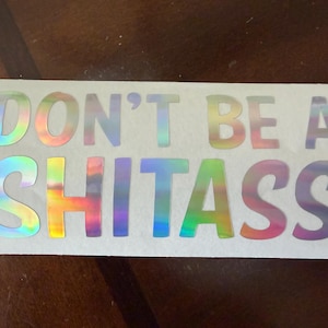 May include: A rectangular sticker with the words "DON'T BE A SHITASS" in a holographic, rainbow-colored font. The text is centered on a white background.