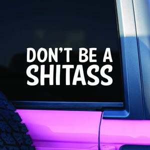 May include: White vinyl decal with the text "DON'T BE A SHITASS" in bold, sans-serif font. The decal is applied to a vehicle window, with a black background and a pink and blue vehicle frame visible.