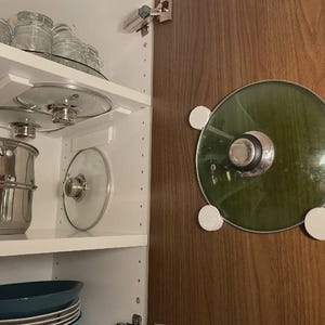 May include: A set of white plastic pot lid organizers mounted on the interior of a wooden cabinet door, holding a circular glass lid with a metal rim and handle. Additional glass lids are stored on white shelves inside the cabinet.