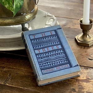 Puede incluir: Un libro antiguo titulado "The Trees" de Conrad Richter, con una cubierta azul, descansa sobre una mesa de madera. Un candelabro de latón con una vela blanca y una jardinera de latón con plantas verdes también están sobre la mesa.