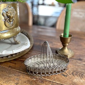 May include: A small, silver wire basket with a handle on a wooden table. The basket is oval and has a woven design. A brass candle holder with a green candle and a brass planter with a lion's head are also visible.