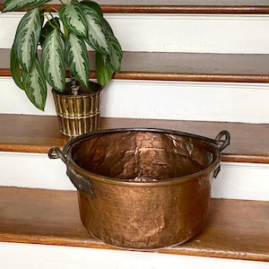 May include: A large, round, hammered copper pot with two dark metal side handles and a visible patina. Beside it sits a green and silver-variegated houseplant housed in a cylindrical, ribbed, metallic gold-toned planter with a vertical pattern.