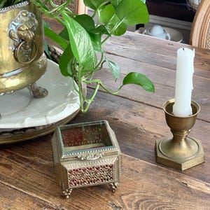 May include: A small rectangular jewelry box with a glass lid, ornate gold-tone metal sides, and red velvet lining, positioned next to a brass candle holder with a white pillar candle. The box features intricate filigree detailing and four feet.