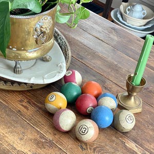 May include: A collection of vintage billiard balls with numbers, in various colours, scattered on a wooden table. A brass candle holder with a green candle and a potted plant are also visible.