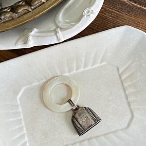 May include: A vintage baby rattle with a silver bell and a white circular handle rests on a white rectangular dish. The bell has decorative detailing and the handle has a pearlescent sheen. The dish is on a wooden surface.