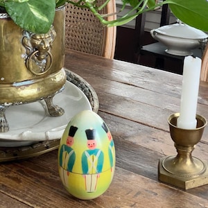 May include: A decorative egg-shaped container with a painted design of toy soldiers in blue and yellow uniforms. The egg sits on a wooden table next to a brass candlestick and a potted plant in a brass planter.