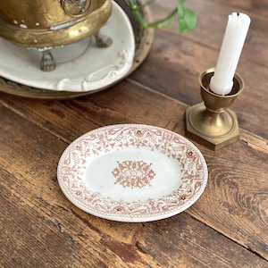 May include: Oval, antique-style plate with a brown floral pattern on a white background. The plate is on a wooden table, next to a brass candle holder with a white candle and a brass planter with a green plant.