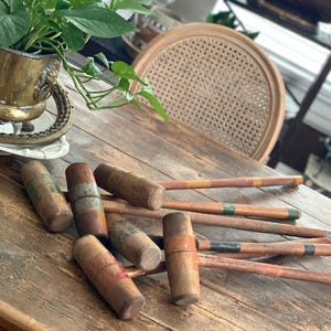 Op de afbeelding: Een verzameling vintage croquet hamers met houten handvatten en gekleurde strepen, rustend op een verweerde houten tafel. De hamers zijn zo gerangschikt dat hun koppen in verschillende richtingen wijzen.