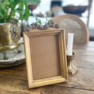 Op de afbeelding: Een goudkleurige rechthoekige fotolijst met een decoratief bloemenaccent aan de bovenkant. De lijst is leeg, met een kartonnen achterkant. Een messing kandelaar met een witte kaars staat rechts van de lijst. De lijst staat op een houten tafel.