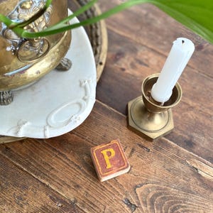 May include: A still life arrangement featuring a brass candle holder with a white candle, a small tile with the letter "P", and a potted plant. The items are on a wooden surface.