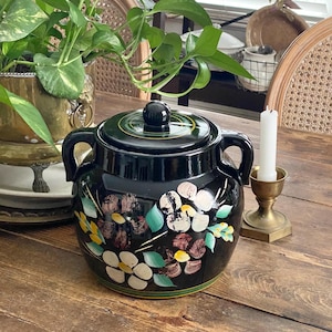 Puede incluir: Un tarro de cerámica negro con tapa y dos asas, adornado con un estampado floral en blanco, amarillo y granate. El tarro está sobre una mesa de madera, con una vela en un portavelas de latón. Se ven hojas verdes en el fondo.