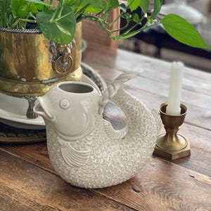 May include: A cream ceramic fish-shaped jug with scale and fin detailing. It rests on a wooden table beside a brass candle holder and a lit white candle. A brass planter with green plants is in the background. The table is made of wood.