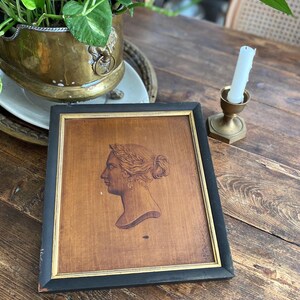 May include: A framed woodcut portrait of a woman in profile, with a laurel wreath and bun hairstyle. The frame is black with a gold inner border. The woodcut is set on a wooden surface.