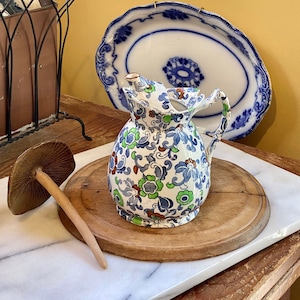 Pode incluir: Uma pequena jarra de cerâmica antiga com um padrão floral em azul, verde e vermelho. A jarra está sobre uma tábua de cortar de madeira, ao lado de um cogumelo. Pratos decorativos azuis e brancos pendurados na parede.