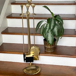 May include: A brass fireplace tool set featuring a stand with a shell-shaped base, a shovel, a poker, and a brush with black bristles. Each tool handle is topped with a decorative brass shell finial. A potted plant sits in a ribbed brass planter.
