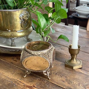 May include: A small, round, footed glass trinket box featuring an intricate gold-tone metal filigree frame. The box has a clear glass lid and sides with a textured, amber-colored base. It stands on four ornate metal legs and includes a decorative metal trim.