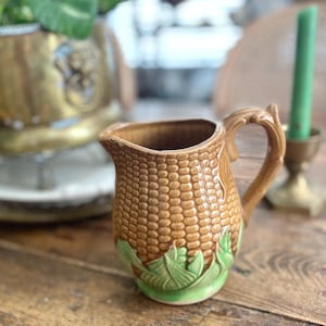Puede incluir: Una jarra de cerámica marrón con un diseño texturizado de mazorca de maíz. La jarra tiene un asa y un diseño de hojas verdes en la base. La jarra está sobre una mesa de madera.