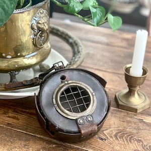 Könnte beinhalten: Ein runder, dunkelbrauner Leder- und Metallgegenstand im Vintage-Stil mit einem messingfarbenen, gitterbedeckten Bullauge. Das Objekt hat Lederriemen und Nieten. Ein Messing-Kerzenhalter mit einer weißen Kerze und ein Messing-Pflanzgefäß mit einem Löwenkopf sind ebenfalls sichtbar.