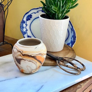 May include: A small, decorative ceramic vase with a marbled pattern in shades of brown, orange, and white. A white hexagonal patterned pot holds a green succulent plant. Vintage scissors and blue and white plates complete the display.