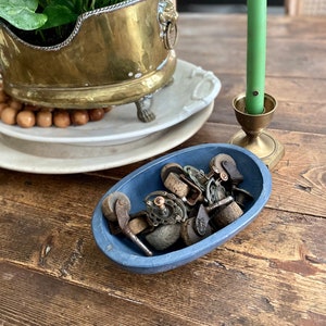 Może przedstawiać: Martwa natura z mosiężną donicą z zieloną rośliną, zieloną świecą w mosiężnym świeczniku i niebieską owalną misą wypełnioną zabytkowymi kółkami. Przedmioty ułożone są na rustykalnym drewnianym stole.