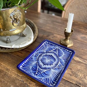 Puede incluir: Una bandeja rectangular de cerámica azul y blanca con un diseño floral. La bandeja está colocada sobre una mesa de madera, junto a una maceta de latón con una cabeza de león y un candelabro con una vela encendida.