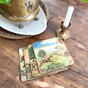 Puede incluir: Juego de cuatro posavasos cuadrados vintage con una ilustración de escena de caza. Los posavasos tienen un borde dorado y representan jinetes a caballo en un paisaje verde y azul. También se ven un candelabro de latón y una planta en maceta.