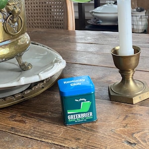 May include: A still life arrangement featuring a vintage brass candle holder with a white candle, a small green and blue tobacco tin labeled "Greenbrier," and a decorative brass planter with greenery. The items are on a wooden table.