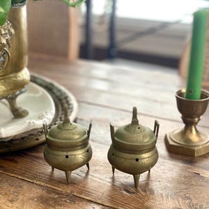 Könnte beinhalten: Zwei kleine, antike Räuchergefäße aus Messing mit Deckeln und drei Beinen stehen auf einem Holztisch. Die Brenner haben ein florales Muster. Eine grüne Kerze in einem Messinghalter ist ebenfalls zu sehen.