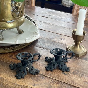 May include: A wooden table displays a brass planter with a lion's head, a white candle in a brass holder, and two black leaf-shaped candle holders. The table has a rustic, aged appearance.