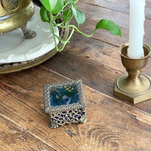 May include: A small square trinket box featuring a dark blue glass base and a decorative gold-toned metal filigree frame. The box has a hinged lid with a clear glass top and sits on four small ornate metal feet, showcasing intricate metalwork.