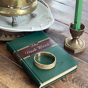 May include: A still life arrangement featuring a green hardcover book titled "The Dark Wood" with a gold-coloured bangle bracelet. A brass candle holder with a green candle and a brass planter with greenery are also present on a wooden surface.