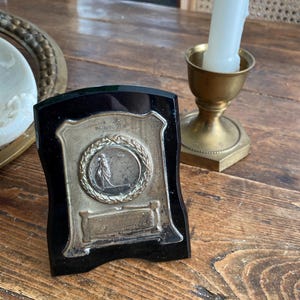 Peut inclure: Plaque vintage noire et argentée en forme de bouclier. La plaque présente un médaillon central avec une figure et une couronne de laurier, et du texte en haut. Un bougeoir en laiton avec une bougie blanche est à droite.
